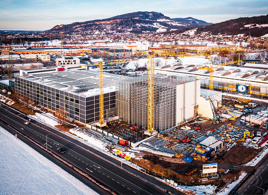 Baustelle neben einer Autobahn mit Kränen und teilweise fertiggestellten Gebäuden vor bergiger Kulisse in der Abenddämmerung.