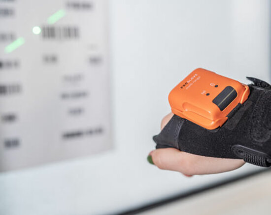Hand trägt einen schwarzen Handschuh mit einem orangefarbenen elektronischen Scangerät und scannt einen Barcode.