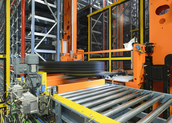                               A tire is just being transferred from the stacker crane to the conveyor system Stacker crane in high-bay warehouse surrounded by stored tires